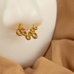 May include: A gold-colored nose ring with a looping, abstract design. The jewelry is displayed on a white surface, set against a backdrop of tan fabric. The nose ring is a unique accessory.