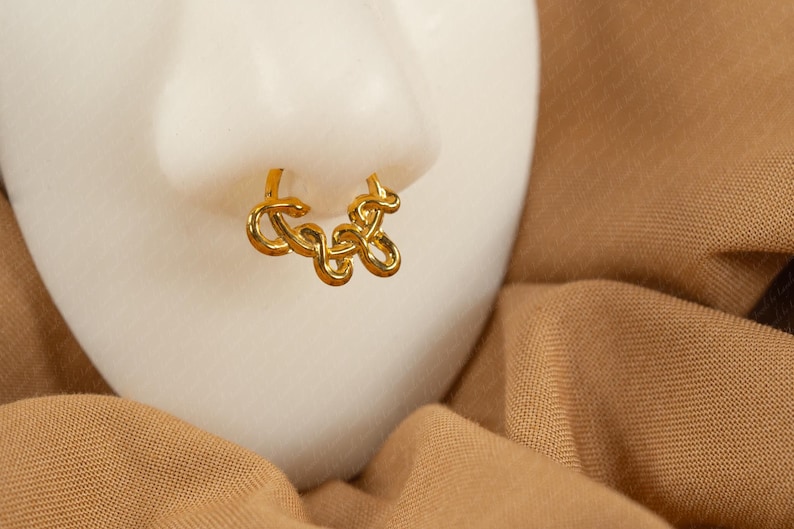 May include: A gold-colored nose ring with a looping, abstract design. The jewelry is displayed on a white surface, set against a backdrop of tan fabric. The nose ring is a unique accessory.