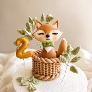 May include: A cake topper featuring a sculpted fox with closed eyes and a green bow tie, sitting inside a tan woven basket. The fox is surrounded by light green leaf sprigs and a gold-colored number two on a clear pick, all on a white base.