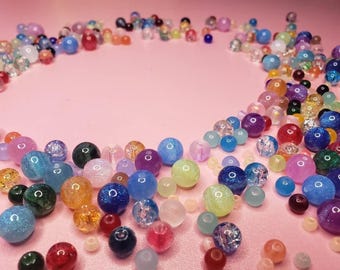 May include: A selection of colourful beads in various sizes and shapes, including round, oval, and irregular. The beads are in shades of blue, green, purple, orange, and pink, with some having glitter or a translucent appearance. They are scattered on a pink surface.
