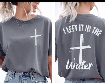 May include: Gray t-shirt with a white cross graphic on the front and back. The back of the shirt features the text "I LEFT IT IN THE WATER" above the cross. The shirt is paired with blue denim jeans.