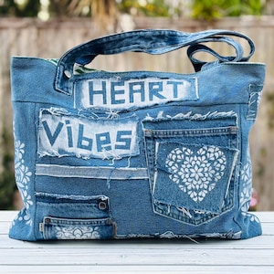 May include: A denim tote bag with the words "HEART" and "VIBES" in white block letters. The bag features multiple denim pockets and a white floral heart design. The bag has denim handles and is made from recycled denim.