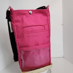 May include: A pink canvas crossbody bag with a clear plastic pocket on the front. The bag has a black adjustable strap and a silver snap closure.