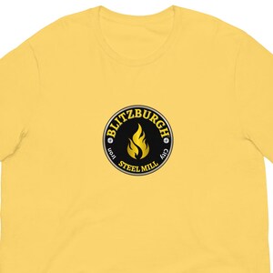 May include: A yellow t-shirt with a circular logo. The logo features the text "BLITZBURGH IRON STEEL MILL CITY" around a flame graphic. The shirt is made of a soft material and has short sleeves.