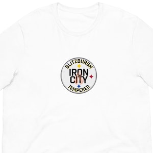 May include: A white t-shirt with a circular graphic that reads "Blitzburgh Iron City Tempered." The graphic includes a star and colour accents. The shirt is made of a soft material and has short sleeves.
