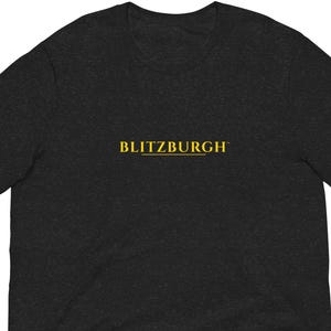 May include: Dark heather gray t-shirt with the word "BLITZBURGH" printed in gold across the chest. The short-sleeved shirt is made of a soft material and has a crew neck.