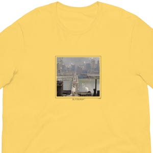 May include: A yellow t-shirt featuring a square image of a bridge and city skyline. The image is in black and white with the word "PITTSBURGH" at the bottom. The t-shirt is made of a soft material.