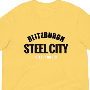 May include: A yellow t-shirt featuring the text "Blitzburgh Steel City" in black, with "Steel Forged" below. The shirt has a crew neck and short sleeves, made from a soft fabric.