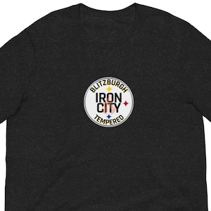 May include: Dark grey t-shirt with a circular graphic. The graphic reads "Blitzburgh Iron City Tempered" in a white circle with a star and colour accents. The shirt is made of a soft material.