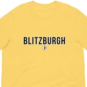 May include: A yellow t-shirt with the word "BLITZBURGH" printed in black across the chest. Below the word is a small circular emblem. The t-shirt is made of a soft material.