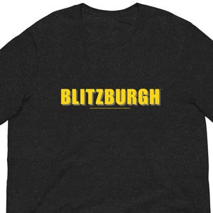 May include: Dark gray t-shirt with the word "BLITZBURGH" in yellow block letters. The text has a shadow effect and is centered on the shirt. The shirt is made of a soft material.