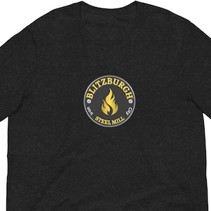 May include: Dark grey t-shirt with a circular logo featuring a flame and the words "BLITZBURGH CITY IRON STEEL MILL" in a yellow and blue design. The shirt is short-sleeved and made of a soft material.