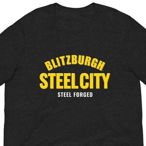 May include: A dark grey t-shirt featuring the yellow text "BLITZBURGH STEEL CITY". Beneath this, in smaller yellow letters, is the phrase "STEEL FORGED". The shirt has short sleeves.