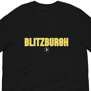 May include: Black t-shirt with the word "BLITZBURGH" in yellow block letters. The word has a black outline. A small circular emblem is centered below the word. The t-shirt is made of a soft material.