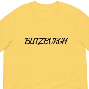 May include: A yellow t-shirt with the word "BLITZBURGH" printed in black cursive across the chest. The shirt is made of a soft material and has short sleeves and a crew neck.