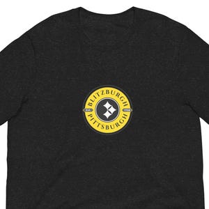 May include: Dark gray t-shirt with a circular logo featuring the text "BLITZBURGH" and "PITTSBURGH" in yellow, surrounding a black and white diamond design. The shirt is short-sleeved and made of a soft material.