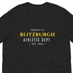 May include: Dark gray t-shirt with the text "PROPERTY OF BLITZBURGH ATHLETIC DEPT. EST. 1933" in yellow and white. The shirt is a short-sleeved crew neck.