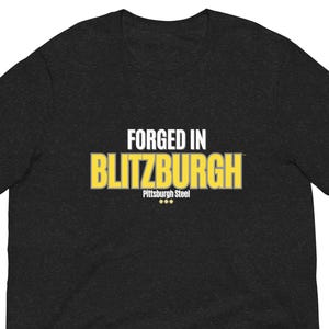 May include: Dark gray t-shirt with the text "FORGED IN BLITZBURGH" in white and yellow block letters. Below the text is "Pittsburgh Steel" in smaller white letters. The shirt is short-sleeved.
