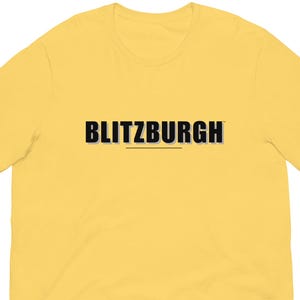May include: A yellow short-sleeve t-shirt with the word "BLITZBURGH" printed across the chest in black, with a thin underline. The shirt is made of a soft material.