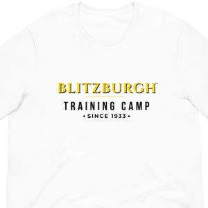 May include: White t-shirt with the text "BLITZBURGH TRAINING CAMP + SINCE 1933+" in yellow and black. The shirt is made of a soft material and has short sleeves.