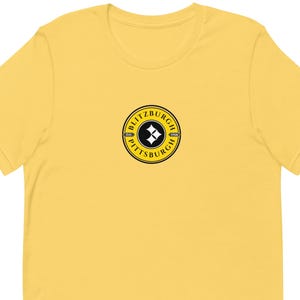 May include: A yellow t-shirt featuring a circular logo with the text "BLITZBURGH" and "PITTSBURGH" in a black and yellow design. The logo includes a black and white diamond shape. The shirt is made of a soft material.