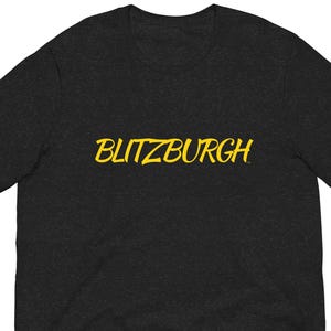 May include: Dark gray t-shirt with the word "BLITZBURGH" in yellow script across the chest. The shirt is made of a soft material and has short sleeves. A casual, everyday wear tee.
