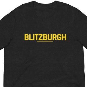 May include: Dark gray t-shirt with the word "BLITZBURGH" in yellow block letters with a yellow underline. The t-shirt is made of a soft material and has short sleeves.