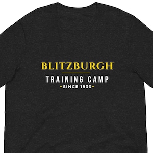 May include: Dark grey t-shirt featuring the text "BLITZBURGH TRAINING CAMP + SINCE 1933+" in yellow and white. The shirt is made of a soft material and has short sleeves.