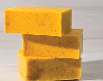 May include: Three stacked, rectangular bars of yellow soap. The soap has a textured surface with small, dark specks throughout. The bars are arranged in a stable, vertical stack, with each bar slightly offset from the one below.