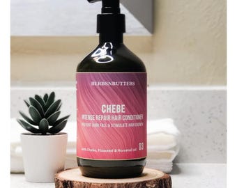 May include: A dark green bottle of CHEBE Intense Repair Hair Conditioner with a black pump dispenser. The label is pink and white with the text "HERBSNBUTTERS" and product information. The bottle sits on a wooden slice with a small succulent plant in a white pot.