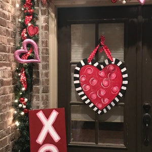 May include: A red wooden sign with white XOXO lettering hangs on a brick wall next to a door decorated with a red and white heart-shaped wreath and a pink heart-shaped door hanger.