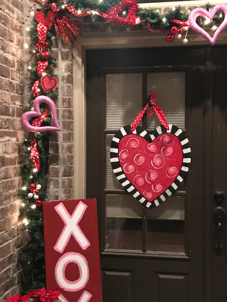 May include: A red wooden sign with white XOXO lettering hangs on a brick wall next to a door decorated with a red and white heart-shaped wreath and a pink heart-shaped door hanger.