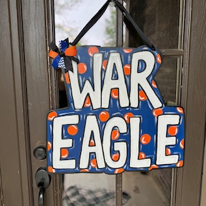 May include: A blue and white wooden door hanger with the words "WAR EAGLE" in white letters. The background is blue with orange polka dots. A black ribbon with a blue and orange bow is attached to the top of the hanger.