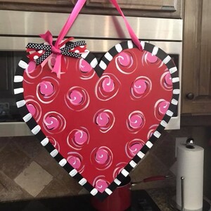 May include: A red heart-shaped door hanger with a white and black striped border. The heart is decorated with pink swirls and a pink bow with black polka dots.