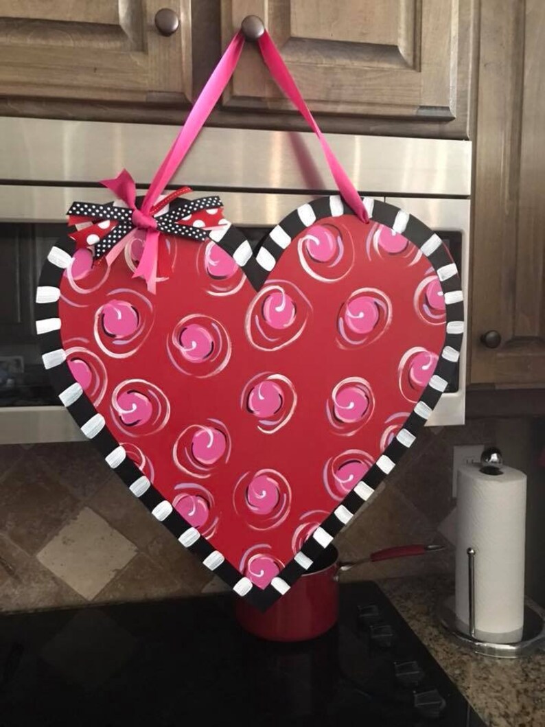 May include: A red heart-shaped door hanger with a white and black striped border. The heart is decorated with pink swirls and a pink bow with black polka dots.