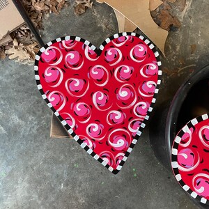 May include: A red heart-shaped wooden sign with a white and black border. The sign is painted with a swirling pink and white pattern.