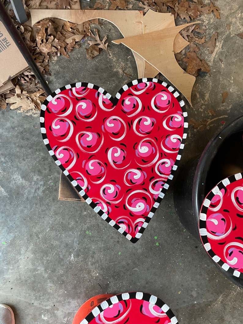 May include: A red heart-shaped wooden sign with a white and black border. The sign is painted with a swirling pink and white pattern.