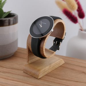 May include: A black Calvin Klein watch with a black leather strap is displayed on a wooden watch stand. The watch has a silver bezel and a black face. The stand is made of light-coloured wood.