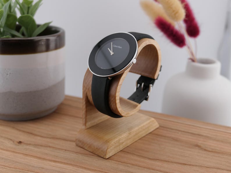 May include: A black Calvin Klein watch with a black leather strap is displayed on a wooden watch stand. The watch has a silver bezel and a black face. The stand is made of light-coloured wood.