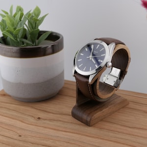 May include: A silver wristwatch with a blue face and brown leather strap is displayed on a wooden watch stand. The watch is presented on a wooden surface, with a small potted plant in the background.