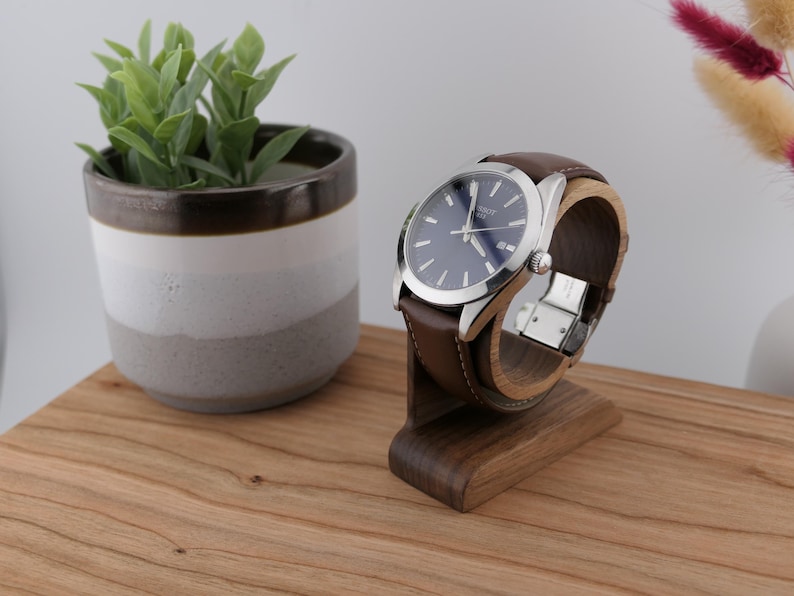 May include: A silver wristwatch with a blue face and brown leather strap is displayed on a wooden watch stand. The watch is presented on a wooden surface, with a small potted plant in the background.