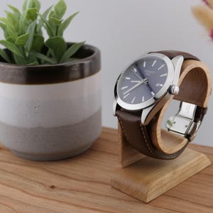 May include: A wristwatch with a blue face and silver casing, featuring a brown leather strap. The watch is displayed on a wooden stand. A small potted plant and decorative flowers are in the background.