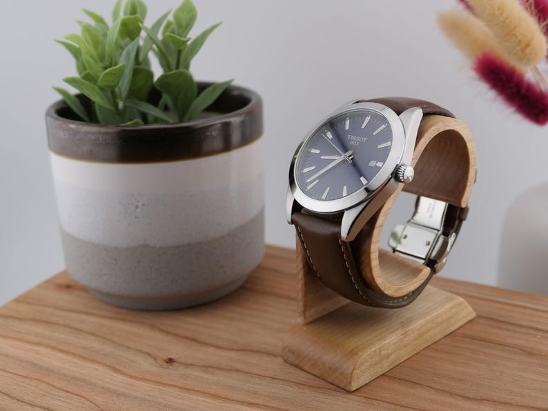 May include: A wristwatch with a blue face and silver casing, featuring a brown leather strap. The watch is displayed on a wooden stand. A small potted plant and decorative flowers are in the background.