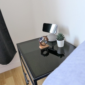 May include: A black glass-topped bedside table with a wooden watch stand, a smart speaker, a small potted succulent, and a phone stand. A wristwatch with a brown leather strap rests on the stand. The scene is set in a bedroom.