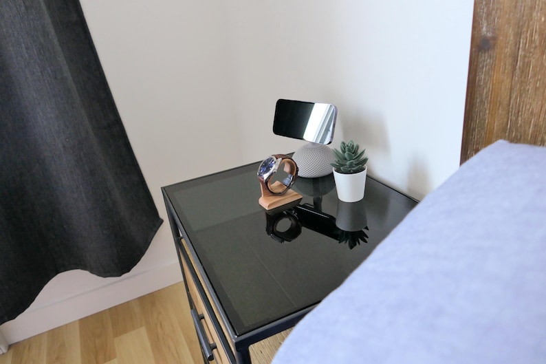 May include: A black glass-topped bedside table with a wooden watch stand, a smart speaker, a small potted succulent, and a phone stand. A wristwatch with a brown leather strap rests on the stand. The scene is set in a bedroom.