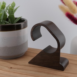 May include: A dark brown wooden watch stand with a curved design. The stand is on a wooden surface, with a potted plant and a vase of dried flowers in the background. The stand is designed to hold a wristwatch.
