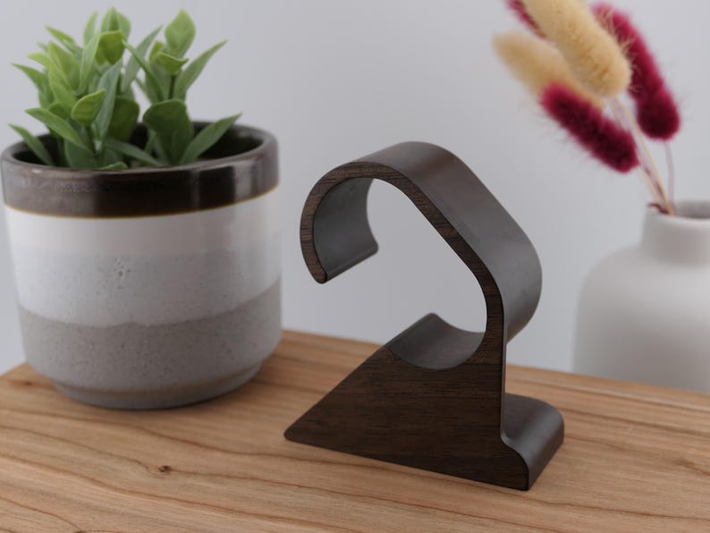 May include: A dark brown wooden watch stand with a curved design. The stand is on a wooden surface, with a potted plant and a vase of dried flowers in the background. The stand is designed to hold a wristwatch.