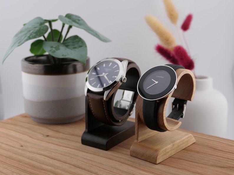 May include: Two wristwatches on wooden stands. One has a blue face and brown leather strap, the other a black face and black leather strap. The watches are on a wooden surface with a potted plant and vase in the background.