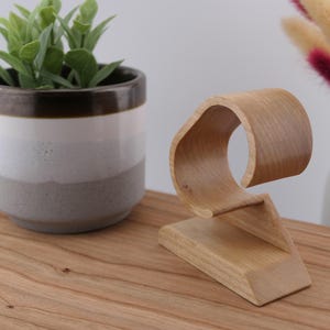 May include: A wooden watch stand with a curved design. The stand is made of light-coloured wood and features a base and a curved top to hold a watch. A small potted plant and decorative flowers are in the background.
