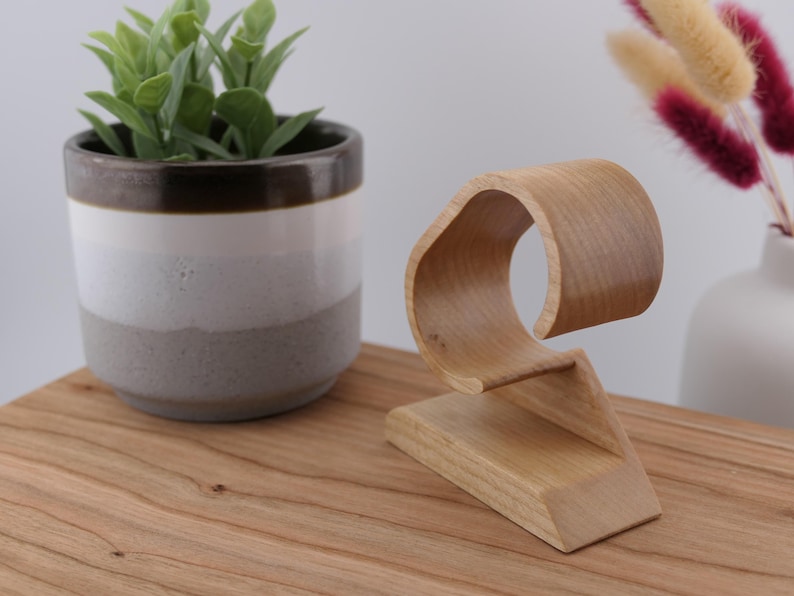 May include: A wooden watch stand with a curved design. The stand is made of light-coloured wood and features a base and a curved top to hold a watch. A small potted plant and decorative flowers are in the background.